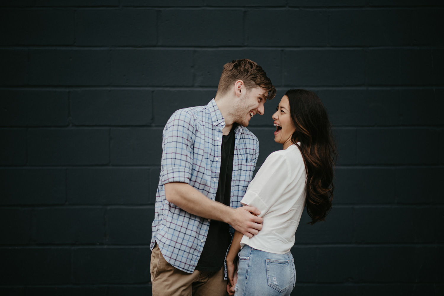Man and woman cuddling and laughing in front of grey cinderblock wall