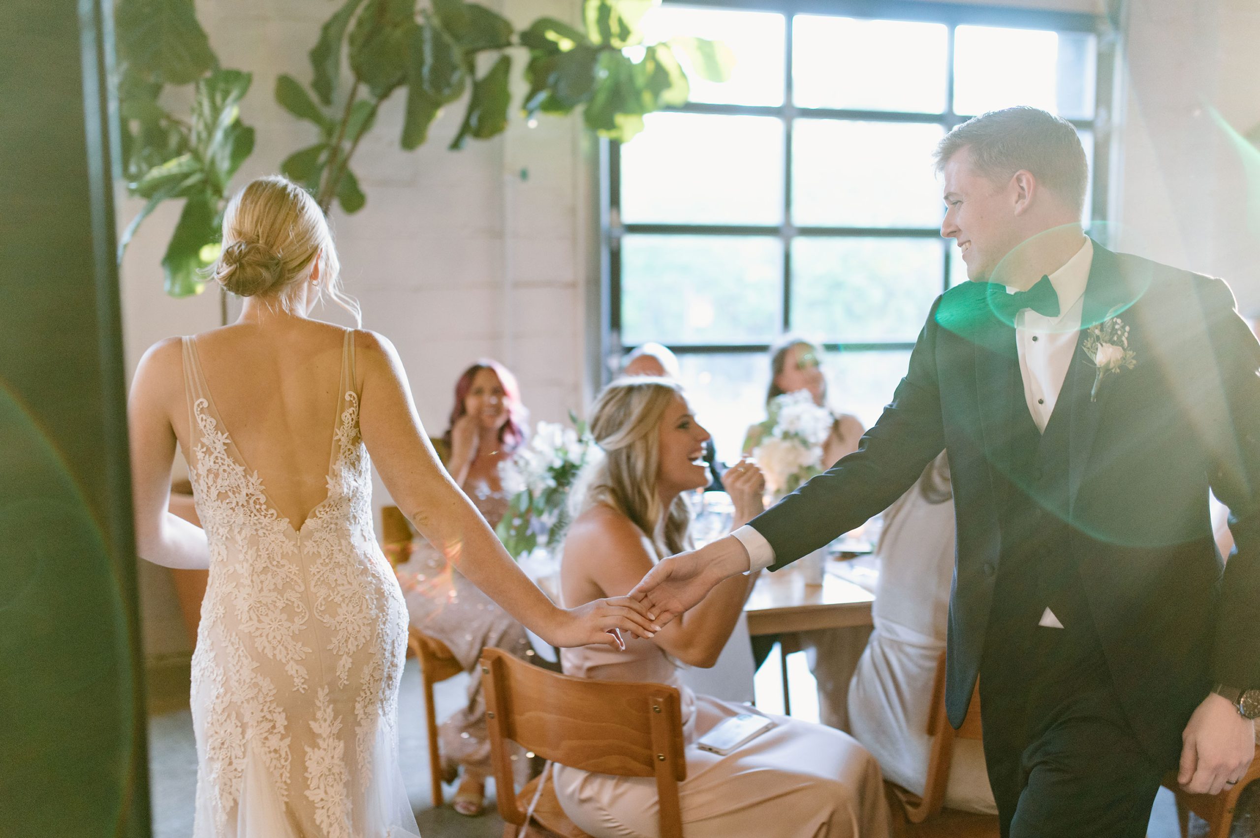 bride and groom hold hands walking through a sunny room towards their table