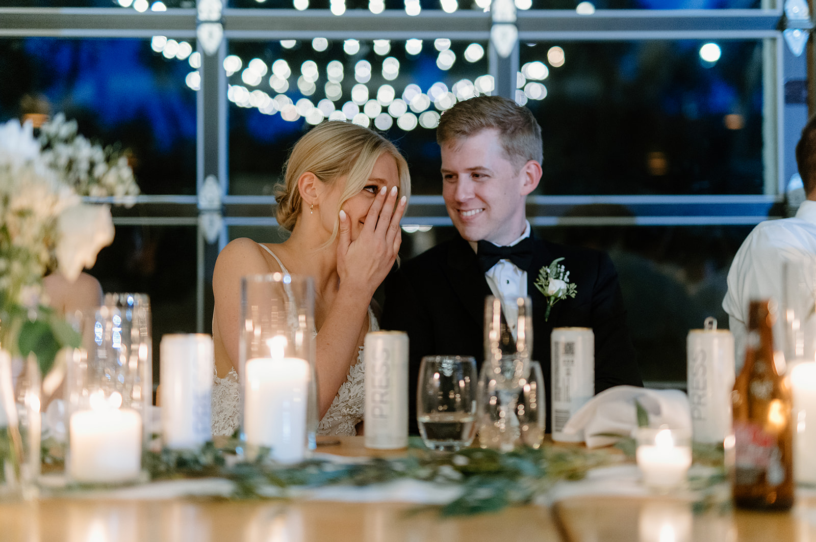 Bride and groom snuggle at reception table surrounded by string light reflection and candlelight
